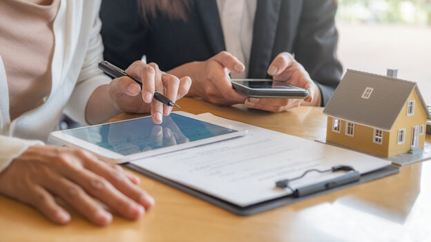 Pessoas analisando contrato de compra de cota contemplada de consórcio com tablet e miniatura de casa sobre a mesa.