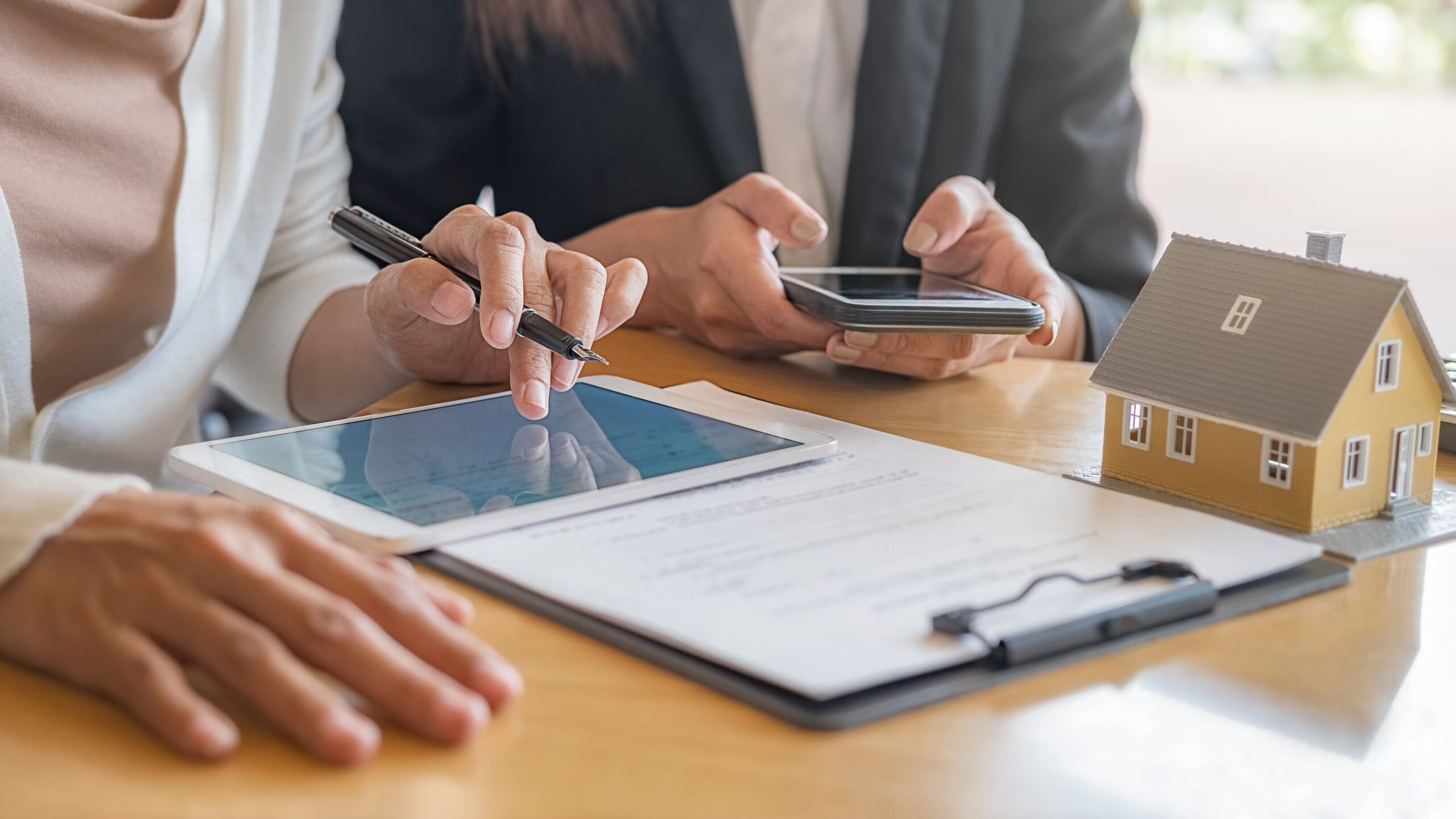 Pessoas analisando contrato de compra de cota contemplada de consórcio com tablet e miniatura de casa sobre a mesa.